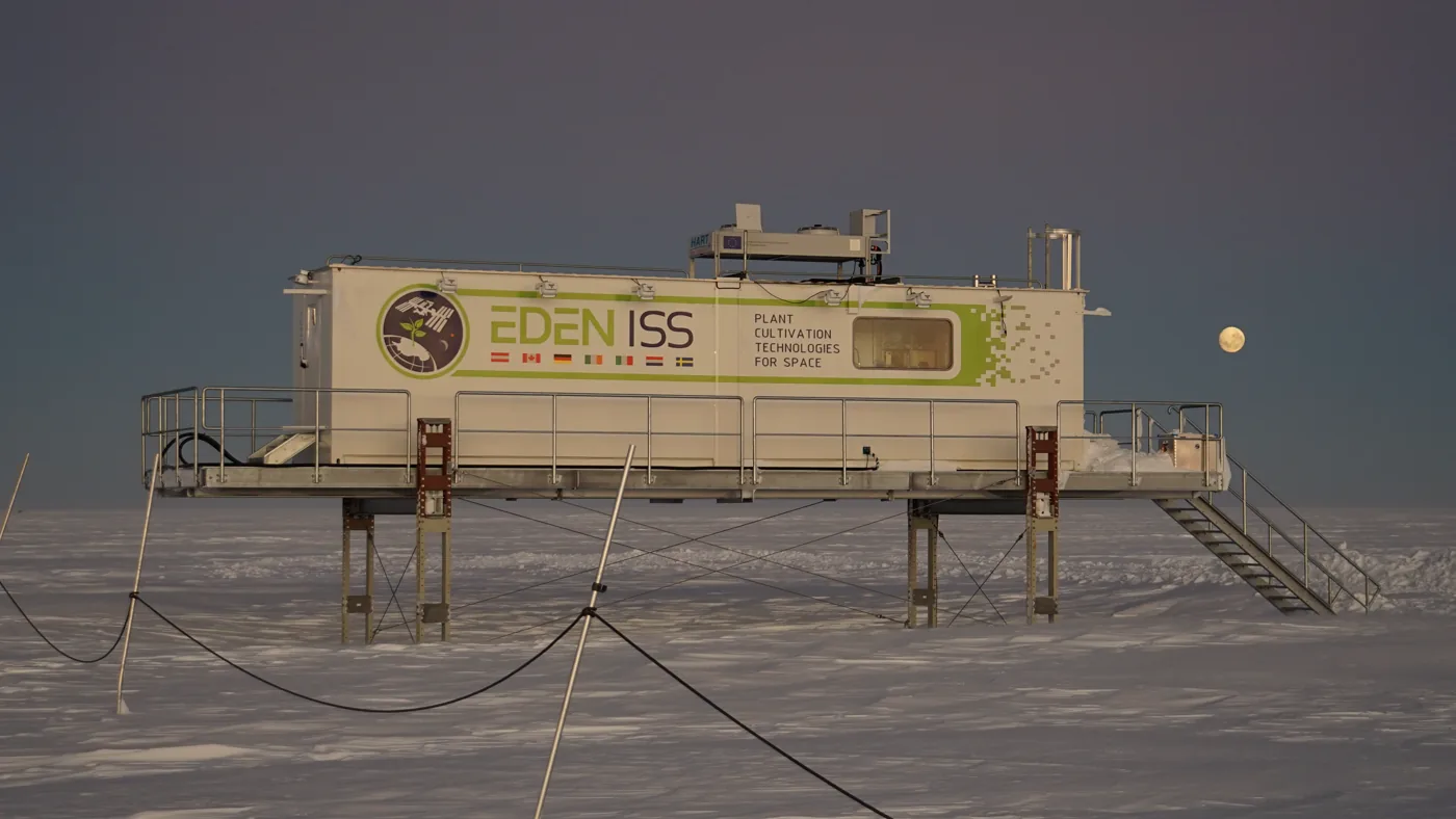 A white container laboratory on stilts above flat Antarctic snow at dusk, a full moon low beside it