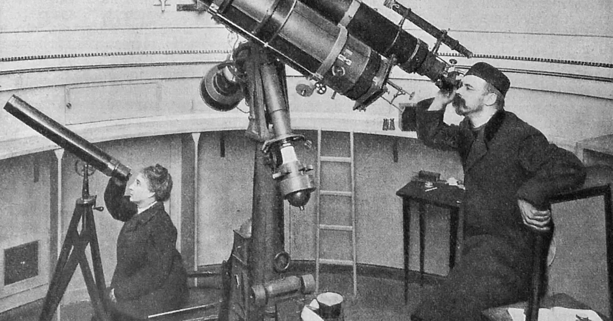 Two people at a large refracting telescope inside an observatory dome, one seated at a smaller guide scope, the other standing at the eyepiece
